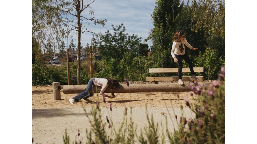 Children playing outside on wooden beams. The sky is blu and you can see plants in the foreground.