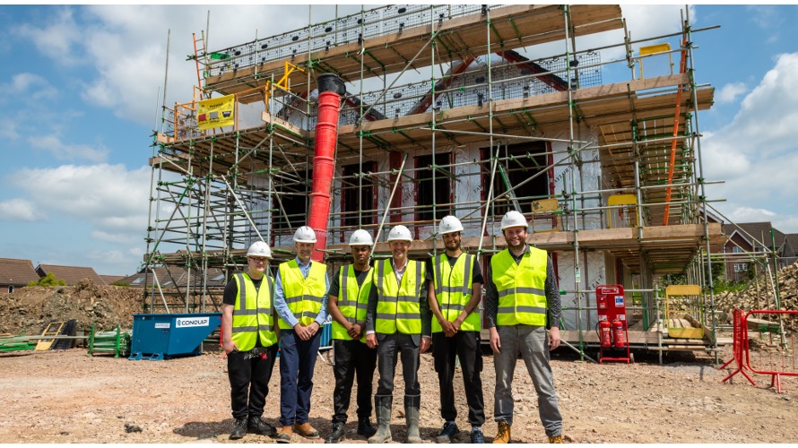 Group of people in hard hats and high vis jackets standing in front of a house that has scaffolding over it, on a building site. Blue sky.