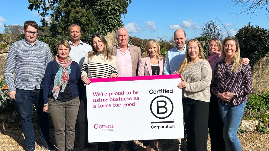 Goram Homes team standing in a group outside on a sunny day. All smiling and holding up a sign that has the B Corp logo on it - and words that say: "We're proud to be using business as a force for good"